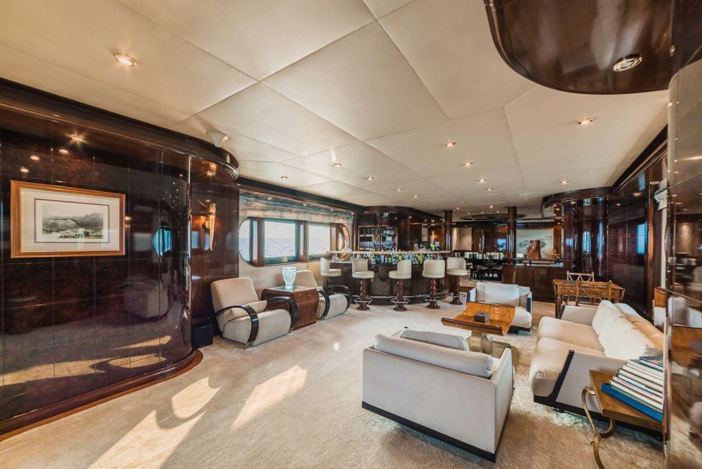 Spacious, luxurious yacht interior with modern dark wood paneling, cream-colored sofas and armchairs, a bar area with high stools, large windows, and contemporary decor. Natural light fills the room.