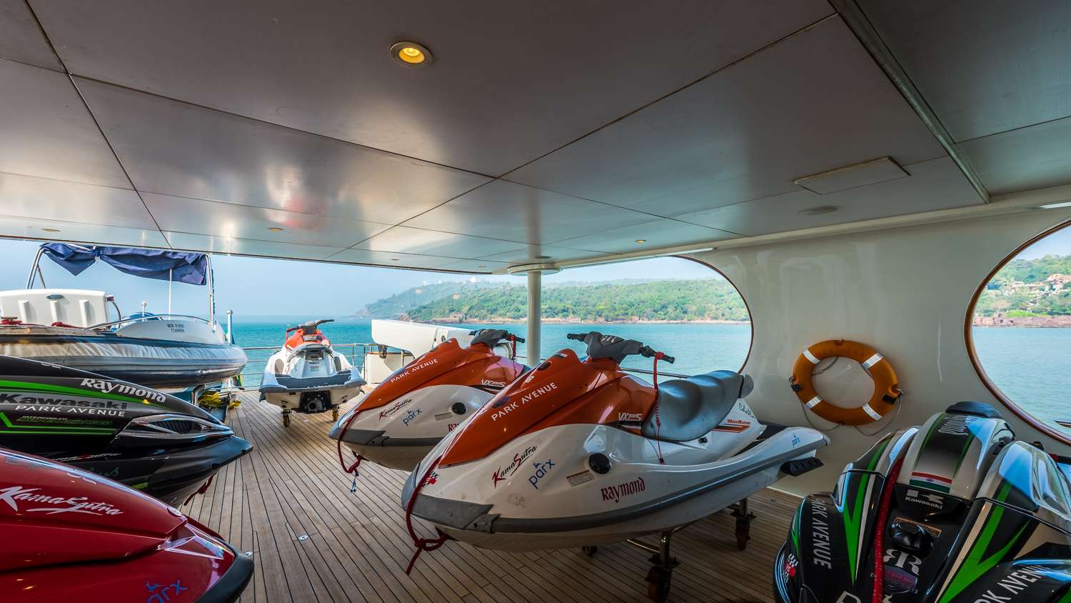 Several jet skis are parked on the wooden deck of a yacht. The yacht overlooks calm blue-green water and a distant shoreline with green hills, visible through large round windows on the side.