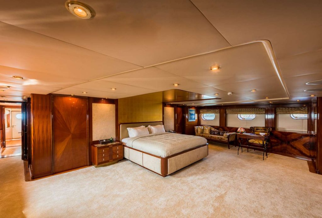 Luxurious yacht bedroom with a large bed, nightstands, a couch by windows, wooden paneling, warm lighting, and beige carpet; hallway visible in the background.