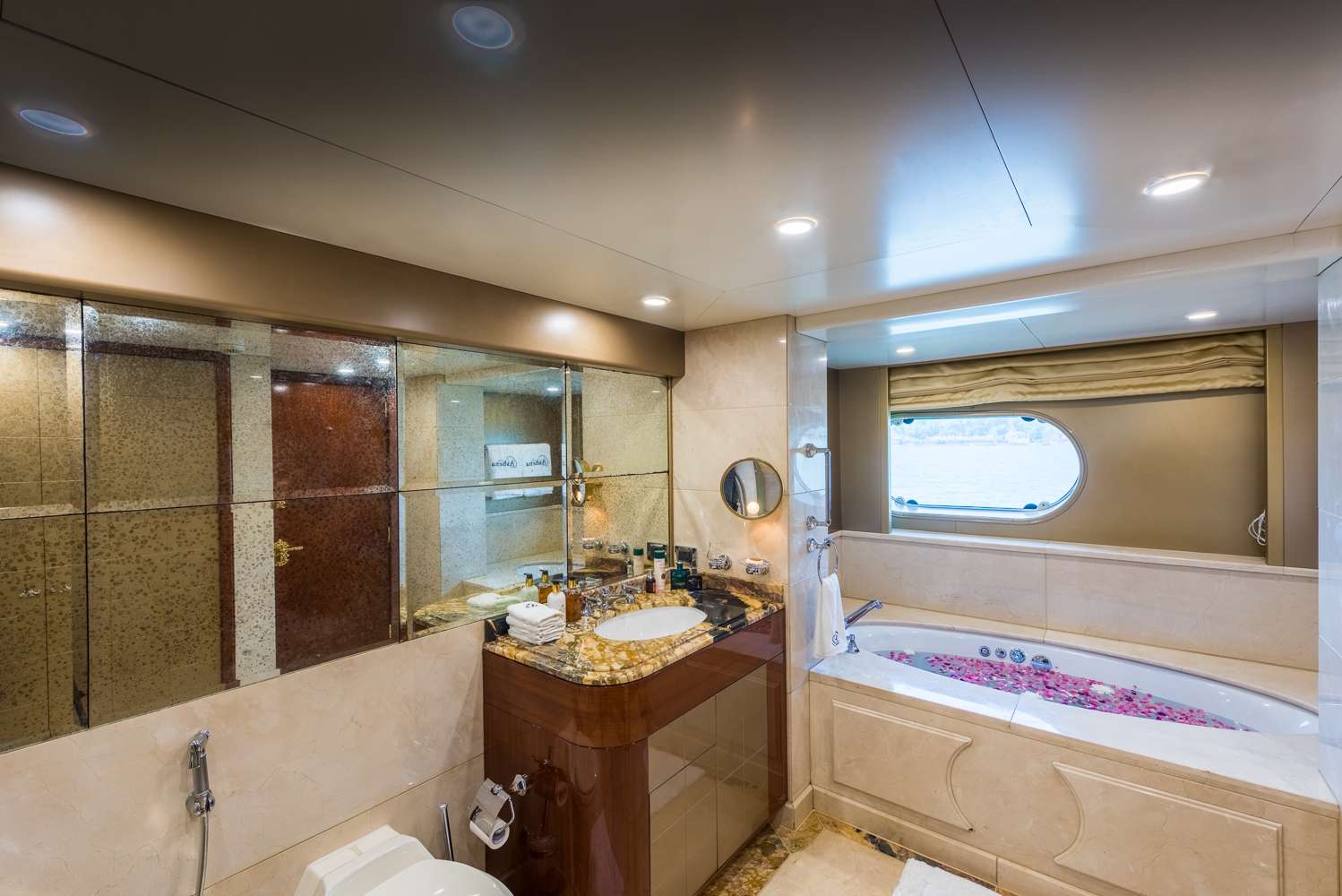 Luxurious bathroom with a large mirror, ornate sink, and counter, a toilet, and a bathtub filled with water and floating flower petals. A window above the tub offers a view of the water outside.