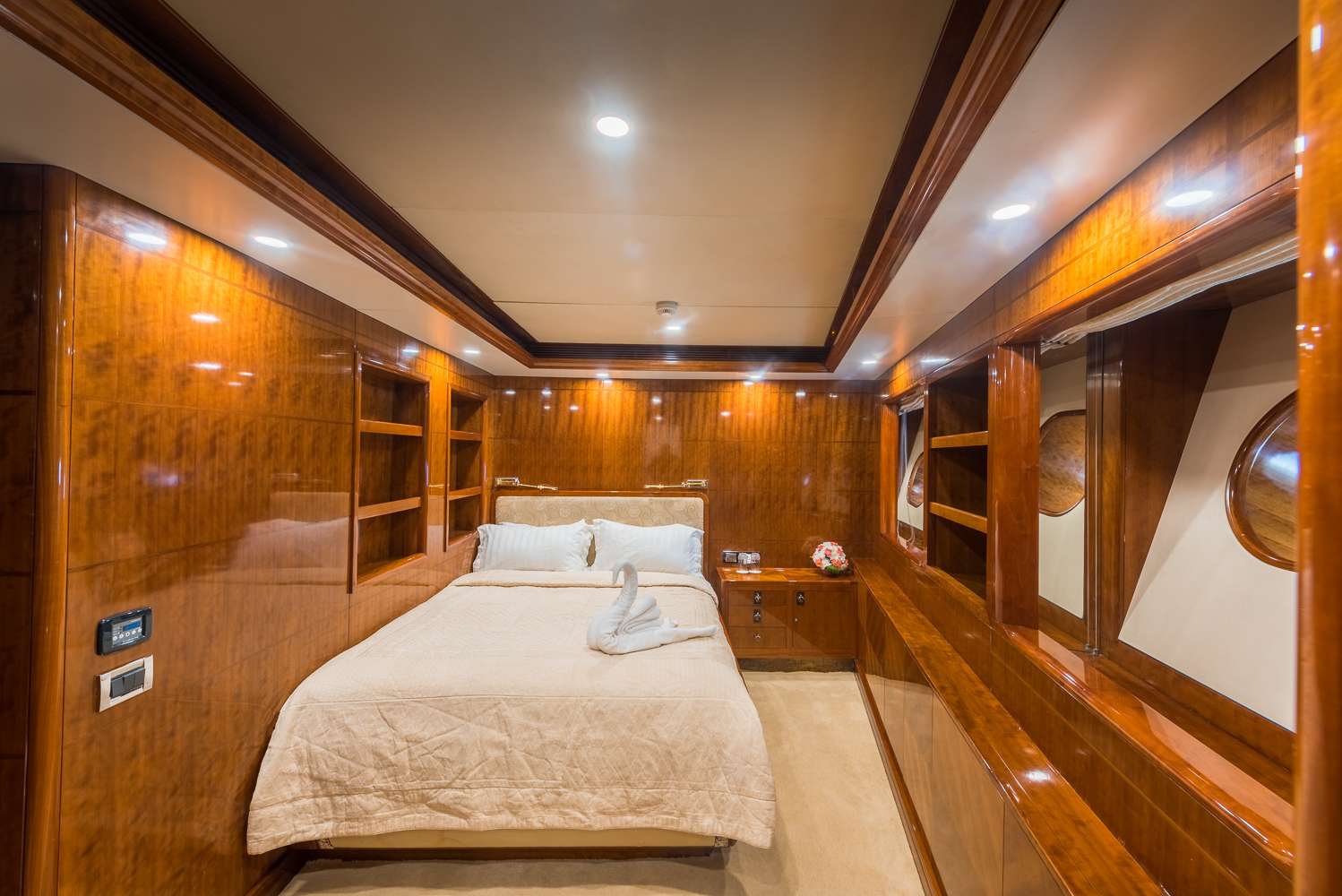A luxurious yacht bedroom with polished wood paneling, a large bed with beige bedding, built-in shelves, mirrored walls, and round porthole windows. Soft lighting creates a warm, inviting atmosphere.