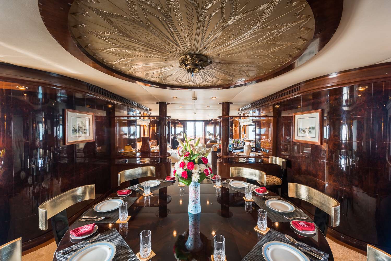 A luxurious dining room with a glossy dark wood table set for eight, elegant chairs, a large floral centerpiece, polished wood-paneled walls, framed art, and an ornate ceiling with intricate detailing.