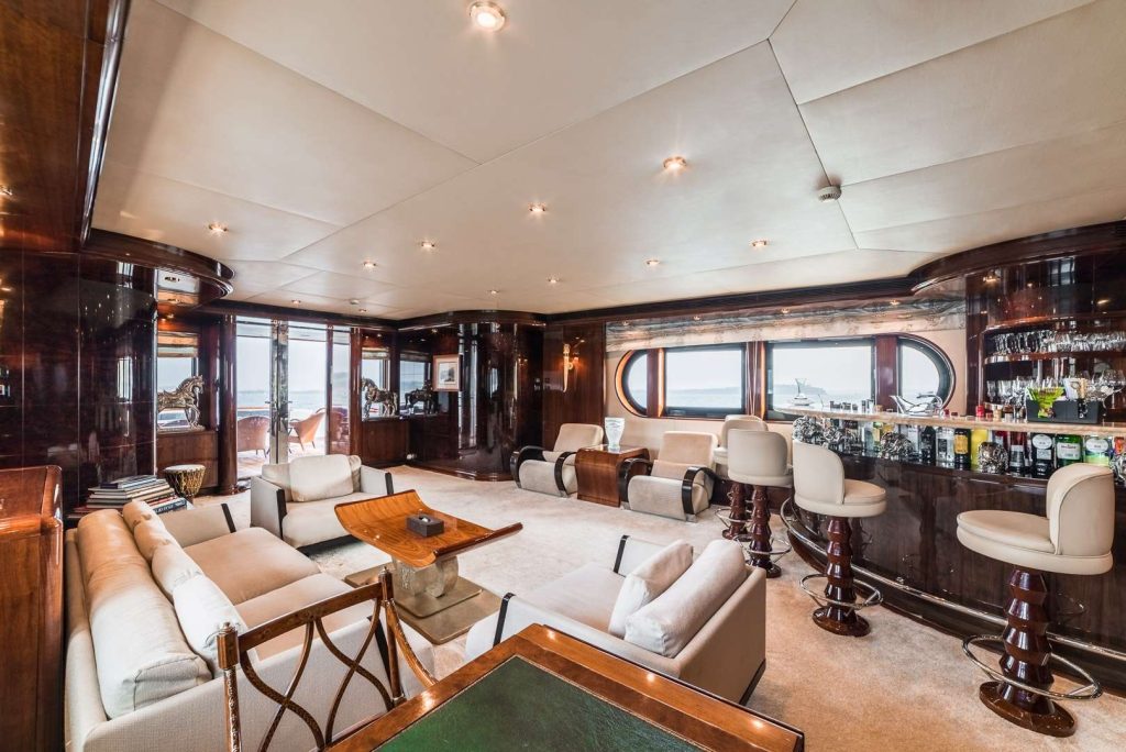 Spacious, luxurious yacht interior featuring cream sofas, a glass-topped bar with stools, polished wood walls, large windows, and soft lighting, offering a comfortable and elegant lounge area with an ocean view.