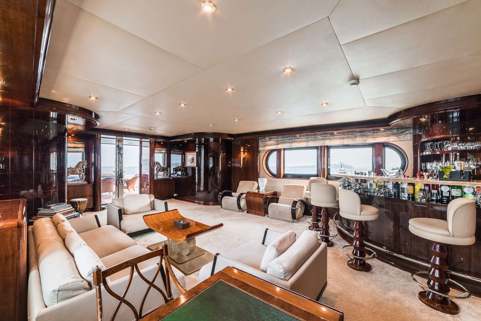 Spacious, luxurious yacht interior featuring cream sofas, a glass-topped bar with stools, polished wood walls, large windows, and soft lighting, offering a comfortable and elegant lounge area with an ocean view.
