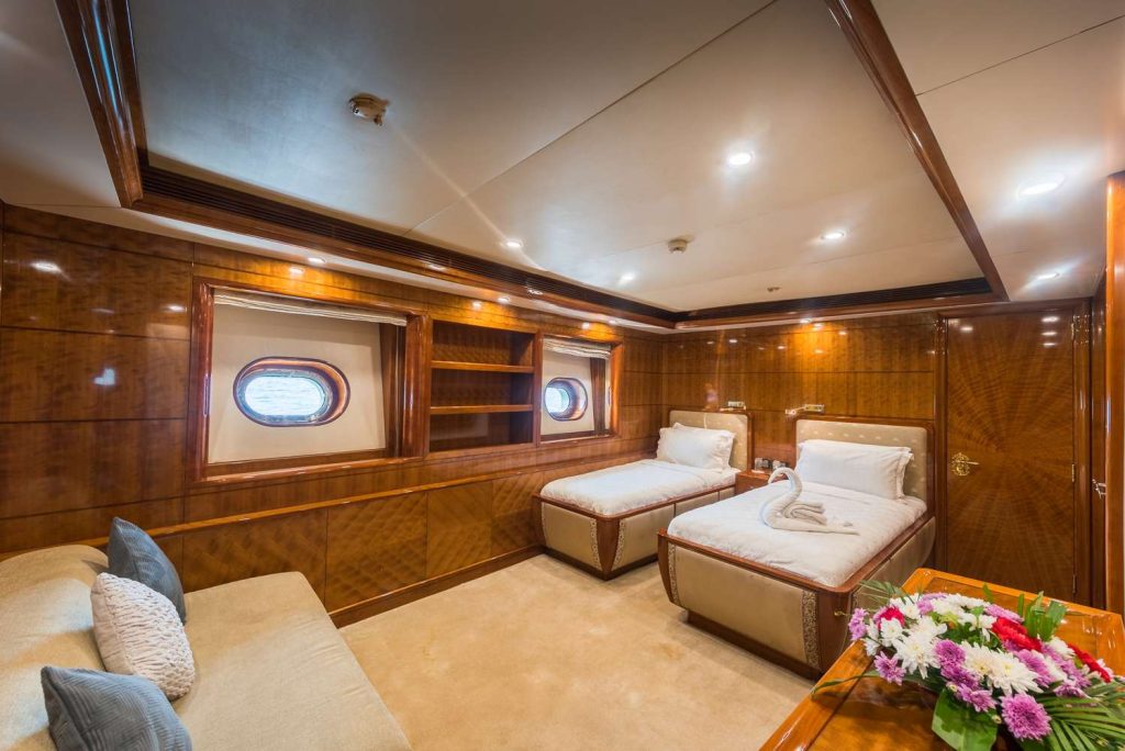 A luxurious yacht cabin with two single beds, wooden paneling, a beige carpet, a sofa with cushions, a flower arrangement on a table, built-in shelves, and two round windows with ocean views.