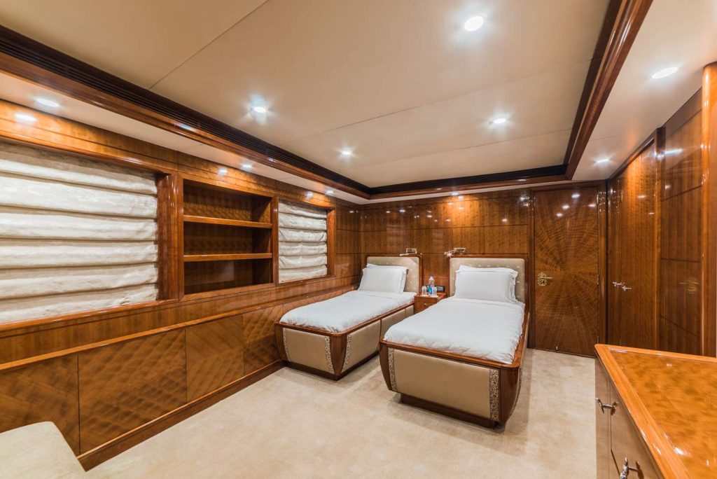 A luxurious yacht bedroom with polished wood paneling, two single beds with white bedding, built-in shelves, and soft overhead lighting, creating a warm and elegant atmosphere.