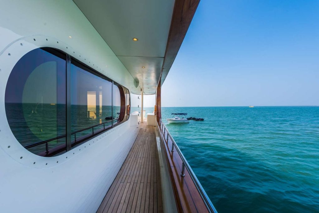 View from the side deck of a modern yacht with wooden flooring, looking out over calm blue ocean water under a clear sky. A small boat floats in the distance.