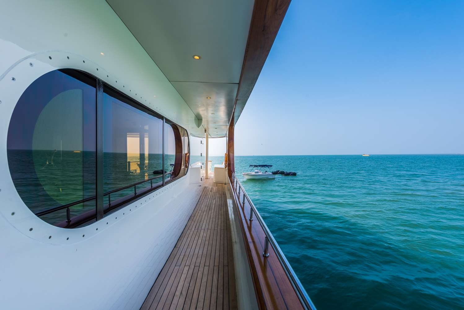 View from the side deck of a modern yacht with wooden flooring, looking out over calm blue ocean water under a clear sky. A small boat floats in the distance.