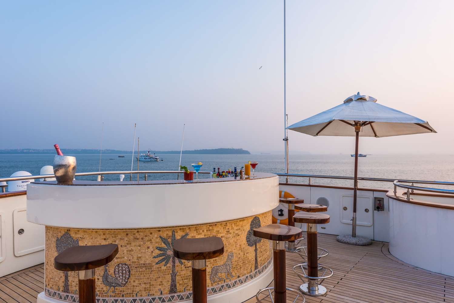 Outdoor bar on a yacht deck with stools, a large umbrella, an ice bucket with a bottle, and assorted drinks, overlooking calm sea waters and distant boats under a clear sky.