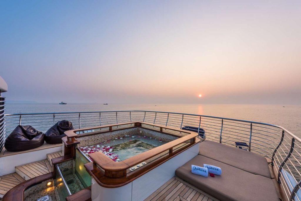 A luxurious yacht deck at sunset, featuring a hot tub with floating flower petals, nearby bean bag chairs, sun loungers, and folded towels, overlooking calm ocean waters.