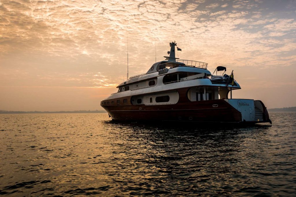 A large yacht floats on calm water at sunset, with a dramatic, cloudy sky overhead and warm orange light reflecting off the surface.