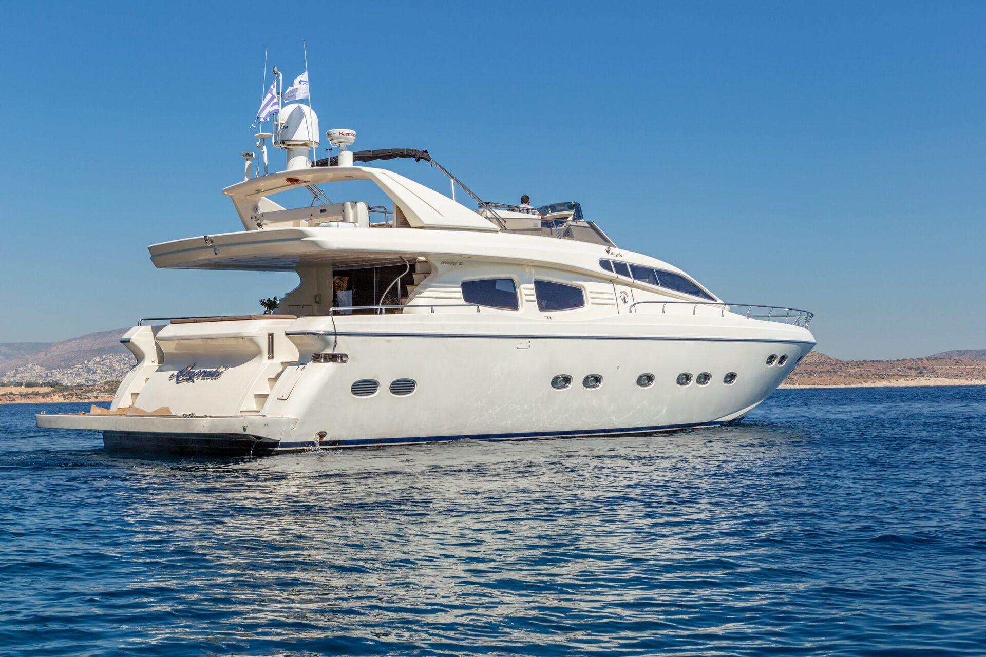 A large white luxury yacht floats on calm blue water near a coastline under a clear, sunny sky. Several flags fly on the yacht, and distant hills are visible in the background.