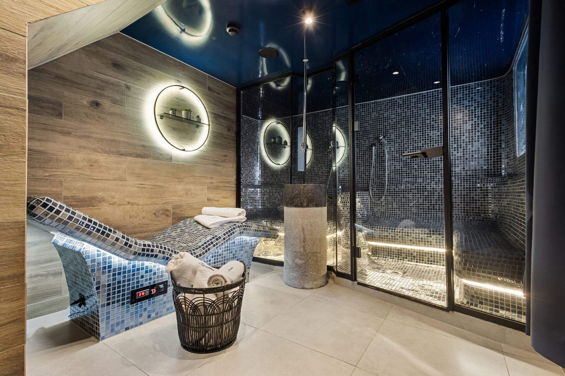 Modern spa room with a mosaic-tiled heated lounge chair, a basket of rolled towels, wood-paneled and tiled walls, and a glass-enclosed steam room with ambient lighting and contemporary decor.