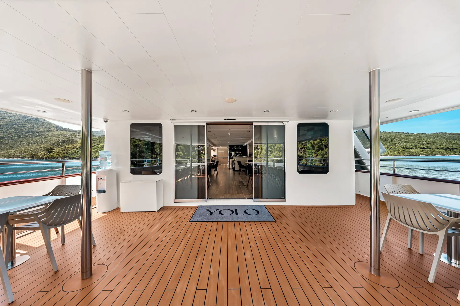Spacious yacht deck with wood flooring, white walls, tables and chairs. Glass doors lead to an interior dining area. YOLO is printed on a doormat. Water and green hills are visible in the background.