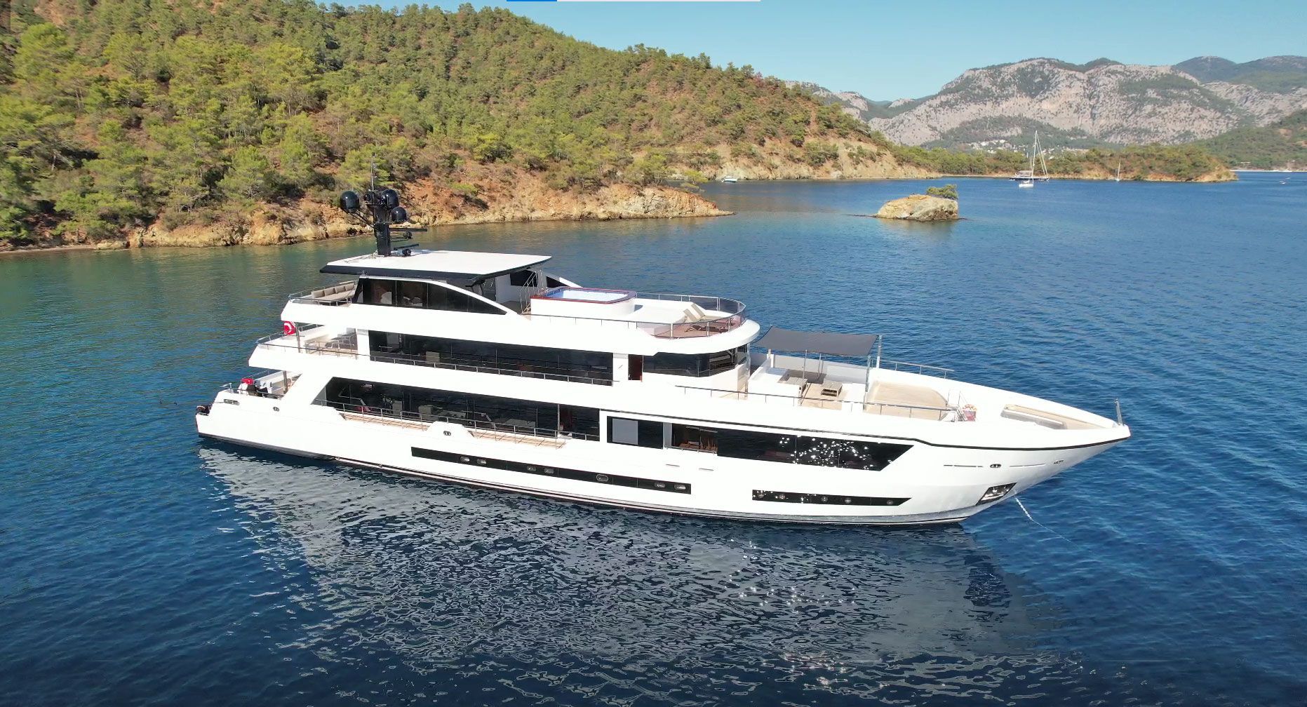 A large white yacht floats on calm blue water near a forested coastline and mountains under a clear sky.