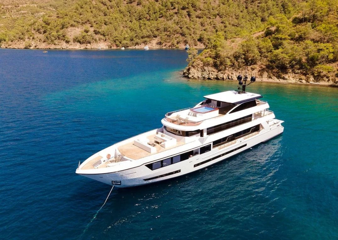 A large white luxury yacht is anchored in clear blue water near a lush, tree-covered coastline with hills in the background.
