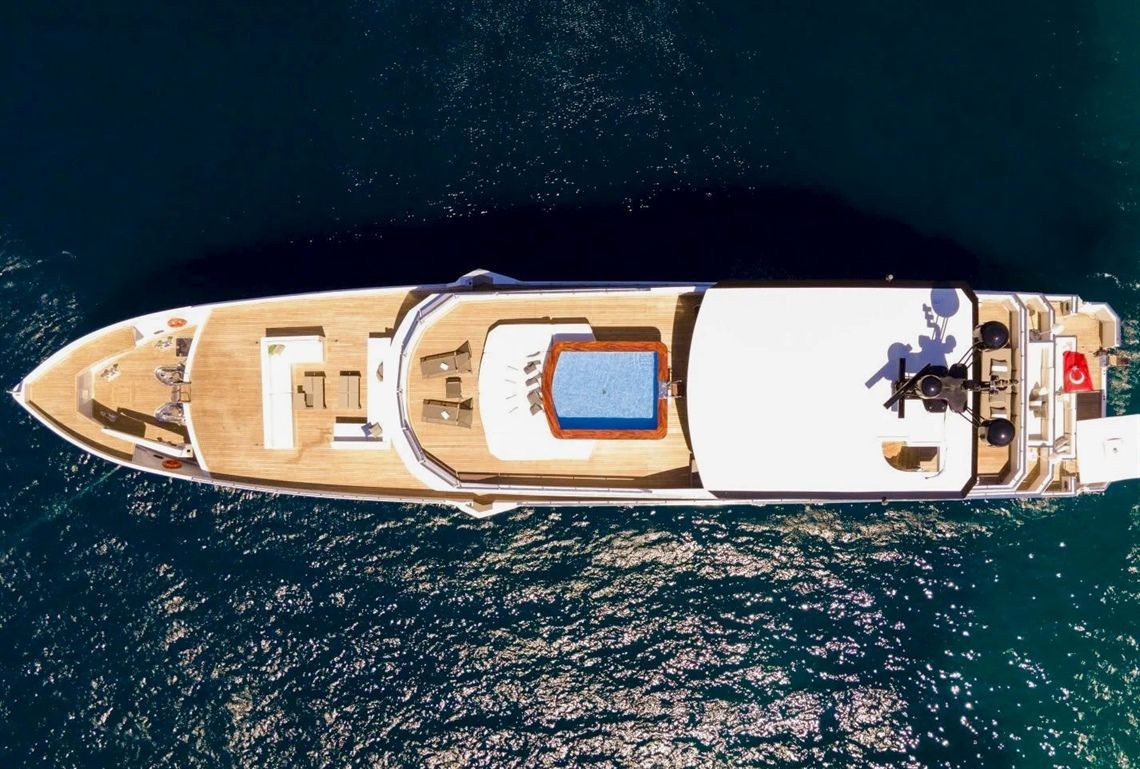 Aerial view of a luxury yacht on deep blue water, showing a sun deck with lounge chairs, a rectangular hot tub, shaded areas, and a visible flag at the stern.