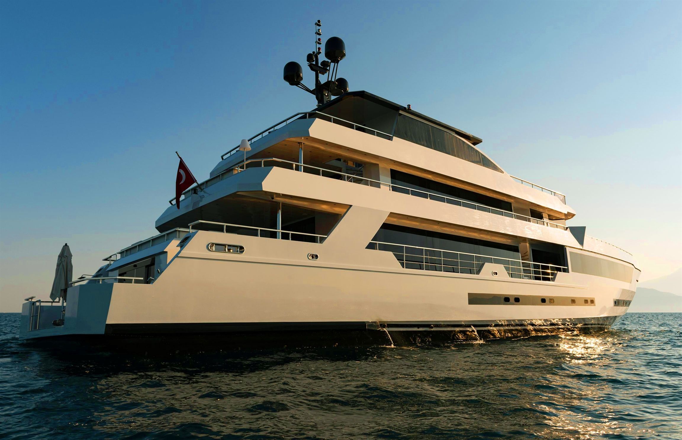 A large, modern white yacht with multiple decks floats on calm ocean water under a clear sky, with a distant shoreline visible on the right.