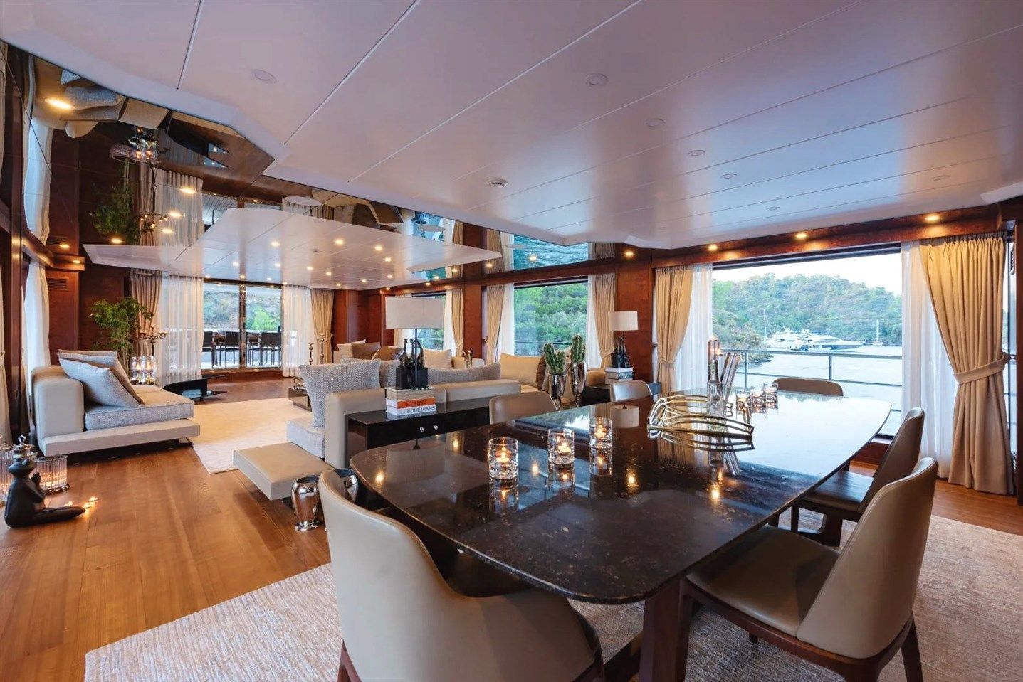 Luxury yacht interior with modern elegant décor, large windows revealing water and green hills outside, plush sofas, a black dining table with beige chairs, and soft ambient lighting.