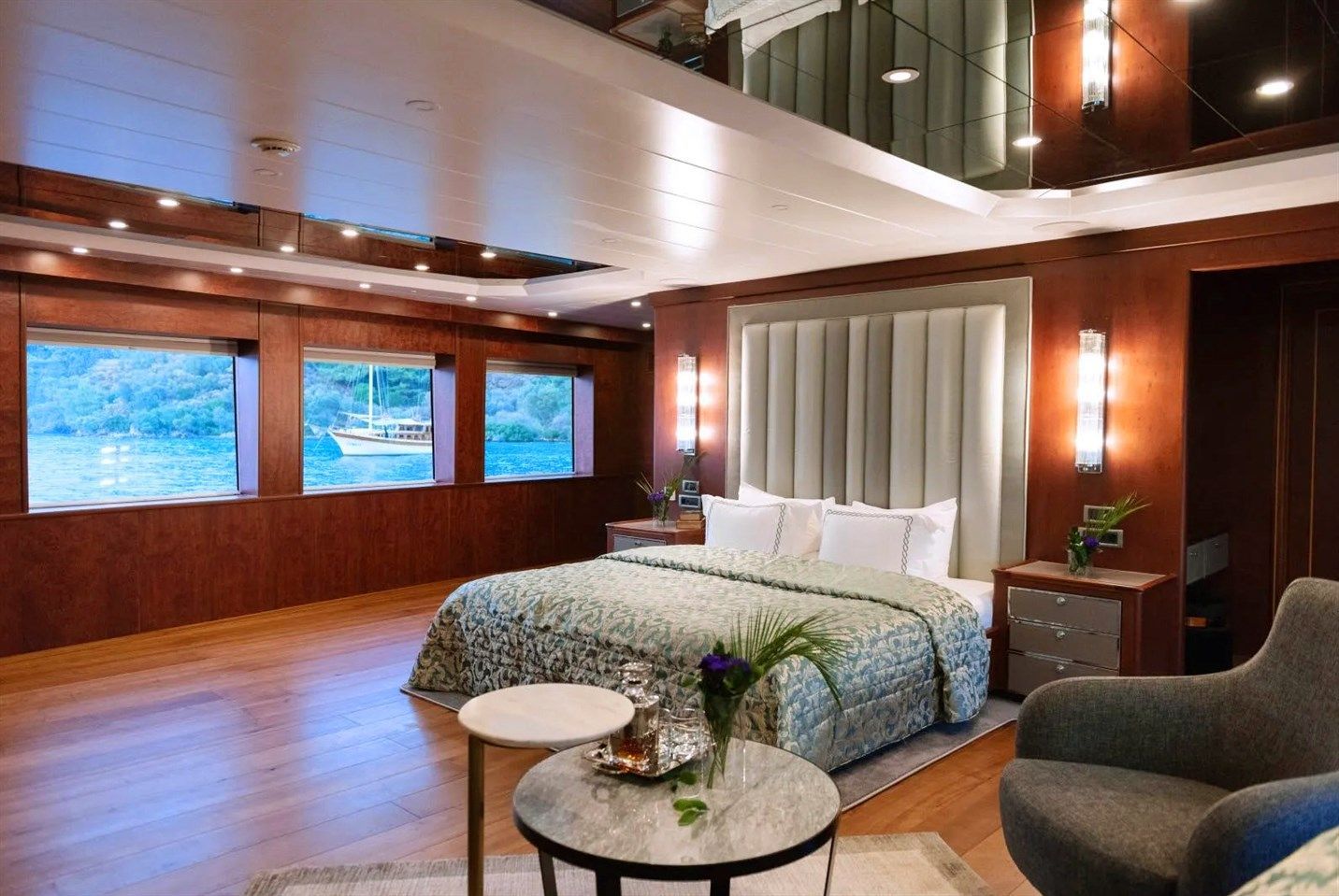 Spacious, modern bedroom with wood-paneled walls, large windows showcasing water and greenery, a bed with a tufted headboard, bedside tables, an armchair, and a small table with flowers and glasses.