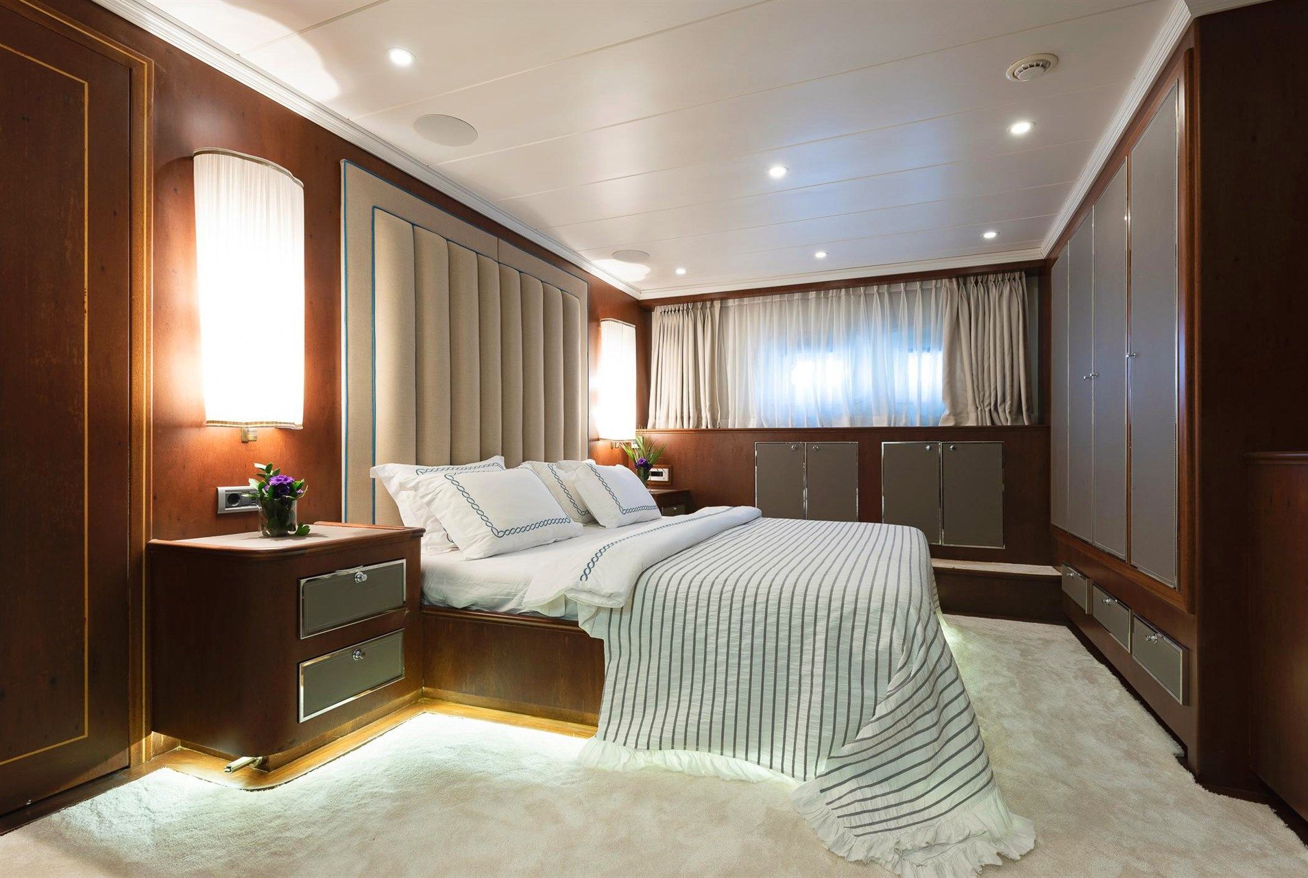 A modern, luxurious bedroom with a large bed, striped bedding, wood-paneled walls, built-in cabinets, soft lighting, and a window with sheer curtains. The room has a calm, elegant atmosphere.
