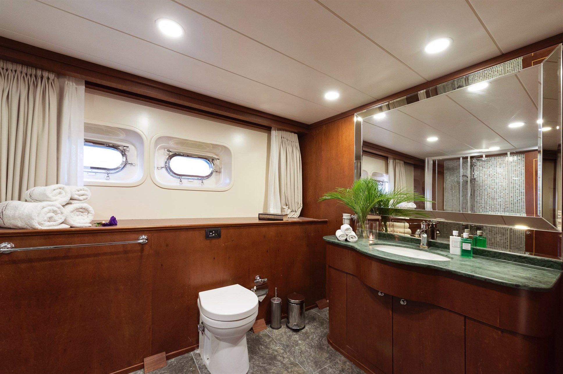 A luxurious yacht bathroom with wood paneling, a toilet, a large mirror above a marble sink, towels neatly stacked by the window, portholes letting in light, and a walk-in shower in the background.