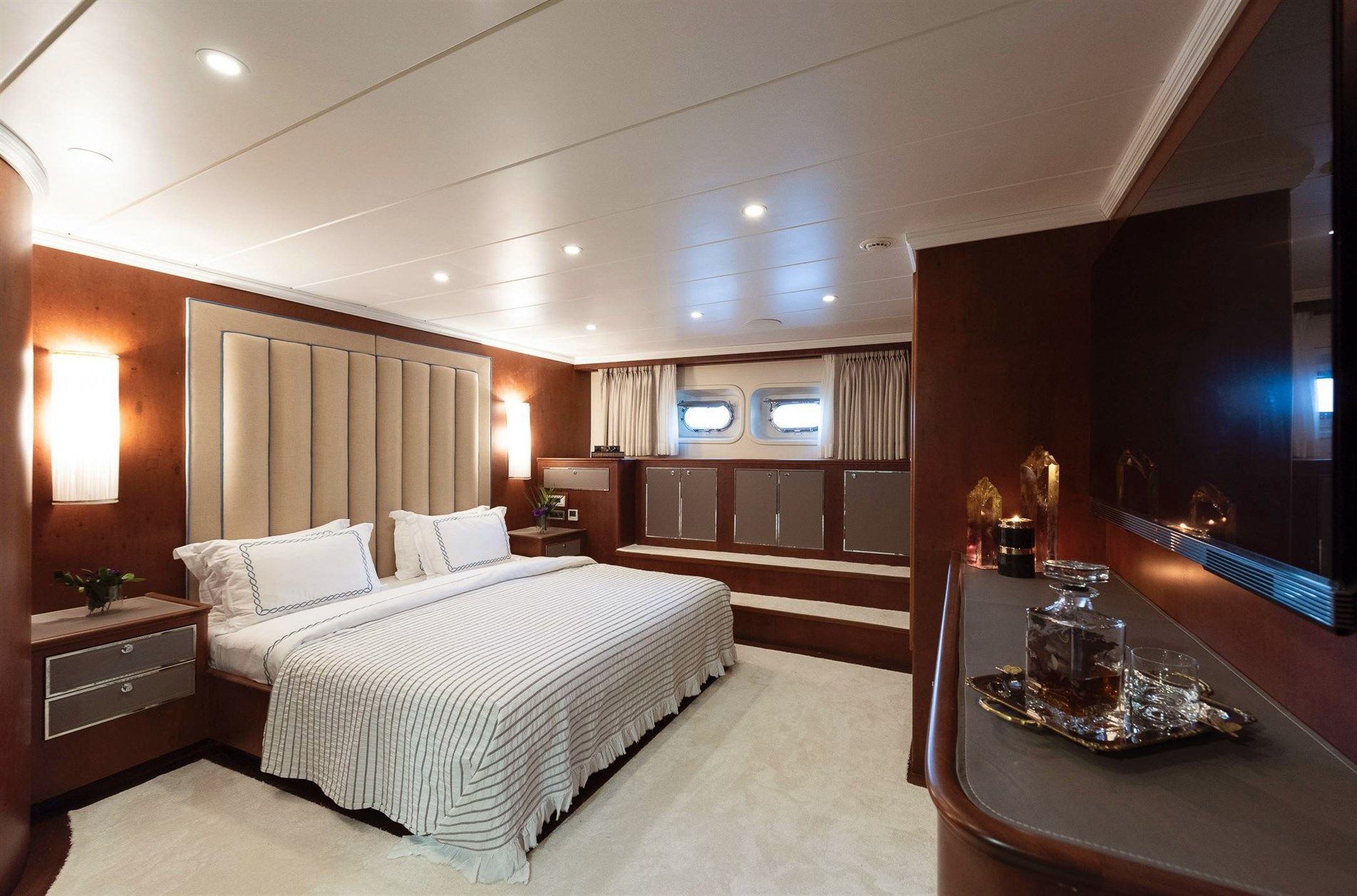 A luxurious yacht bedroom with a large bed, elegant bedding, wood paneling, soft lighting, windows with curtains, and a sideboard with a tray holding a decanter and glasses.