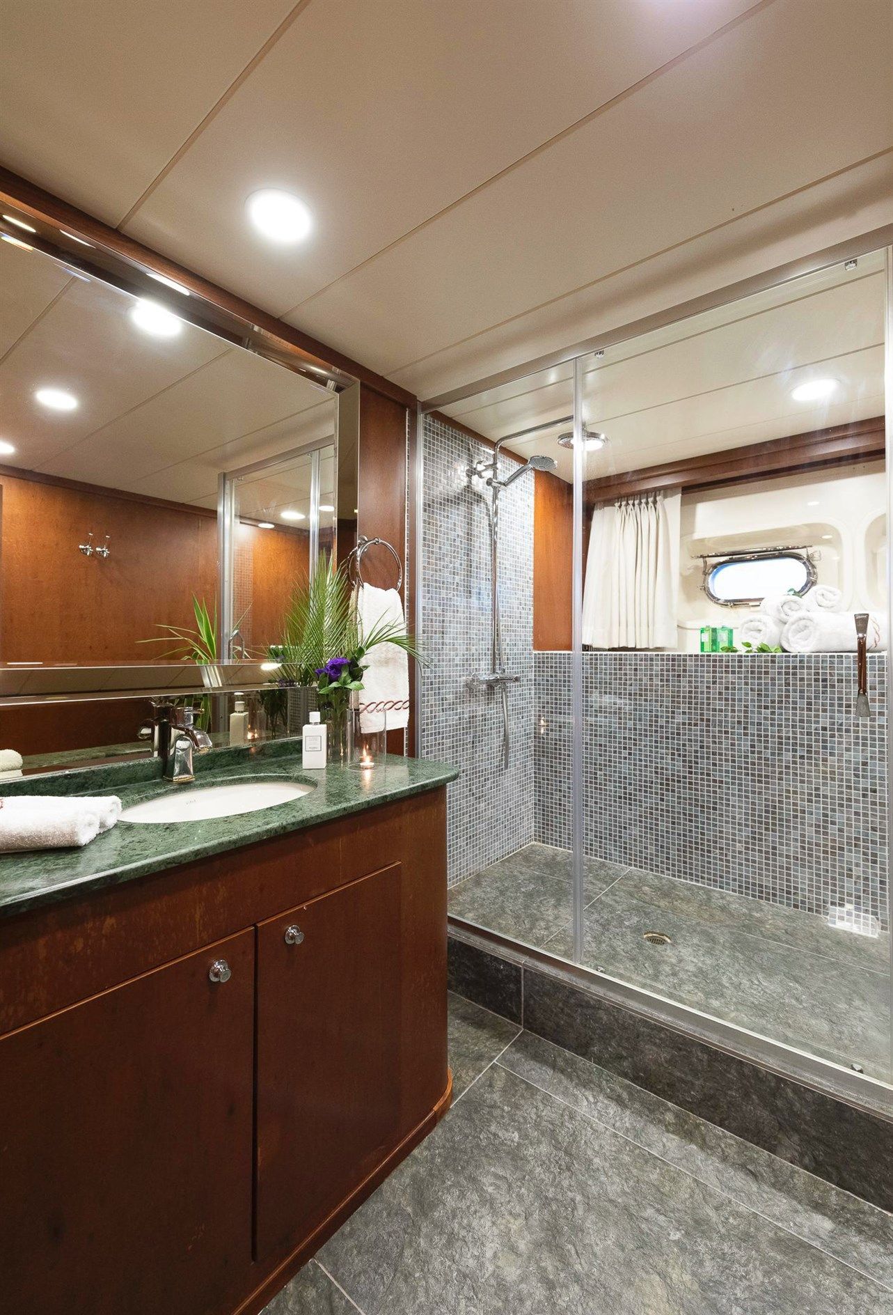 A modern bathroom with dark wood cabinets, a green marble countertop, a large glass-enclosed shower with mosaic tile, a sink with a mirror, towels, and a small window with a curtain.