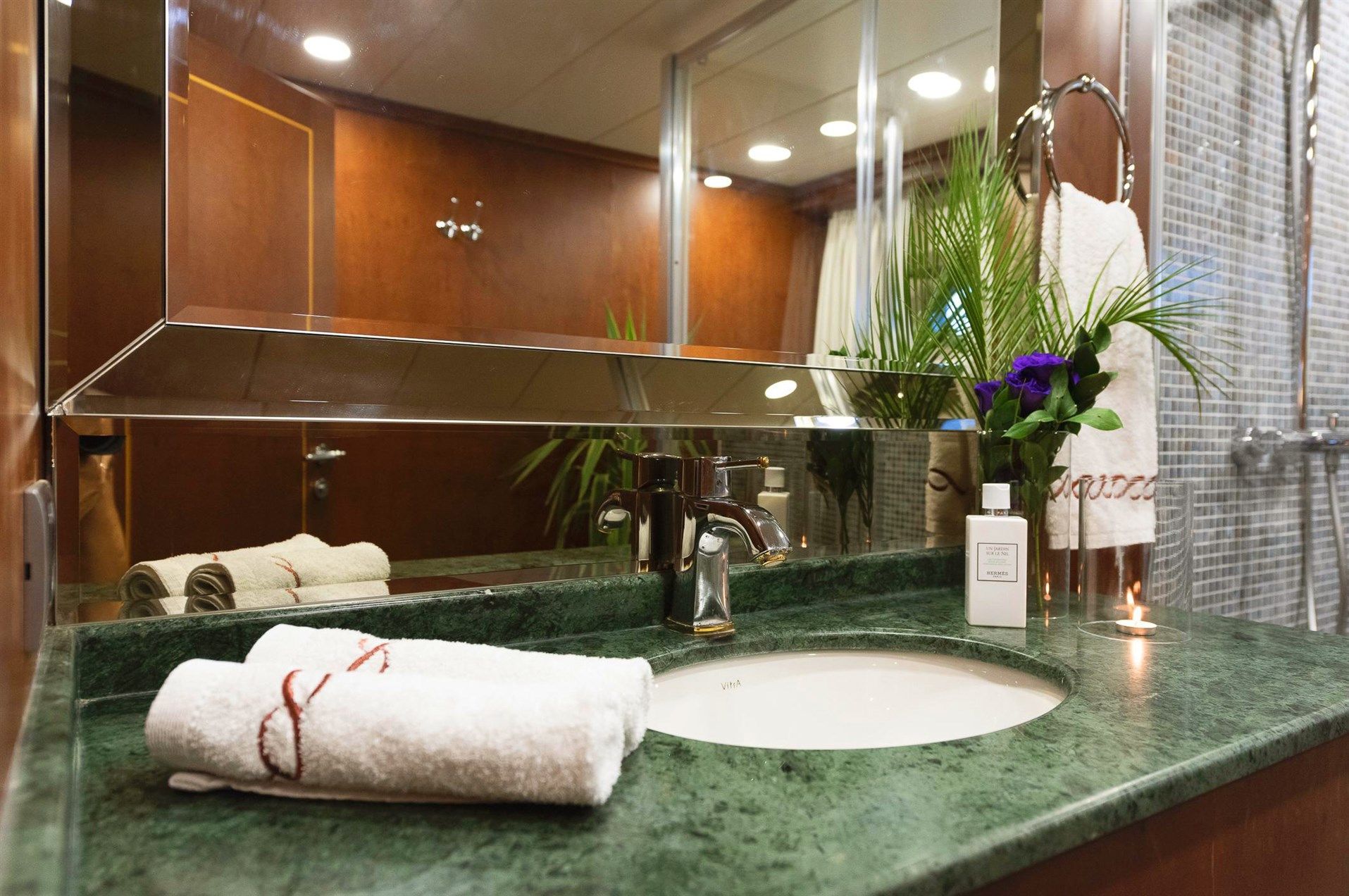 A bathroom with a green marble countertop, white towels, a silver faucet, soap, a vase with purple flowers, and a mirror. In the background are a shower area, plant, and wood-paneled walls.