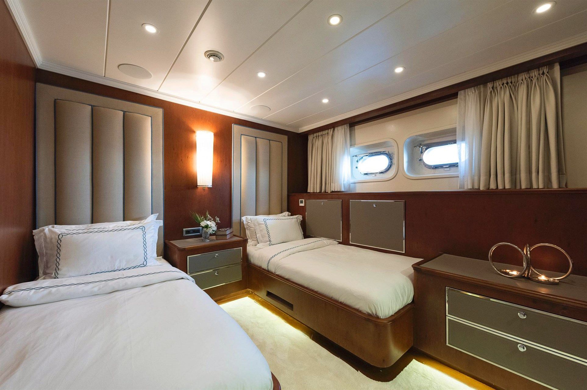 Luxurious cabin with two single beds, modern wood paneling, soft lighting, porthole windows with curtains, and sleek nightstands featuring minimalistic decor and metallic lamps.