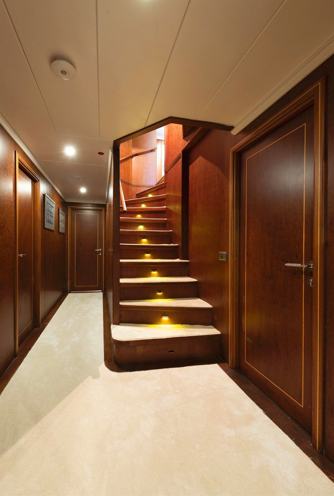 Wood-paneled hallway with beige carpet, two closed doors on either side, and a central staircase with step lights leading upwards to a brighter area. The space has a warm, elegant feel.