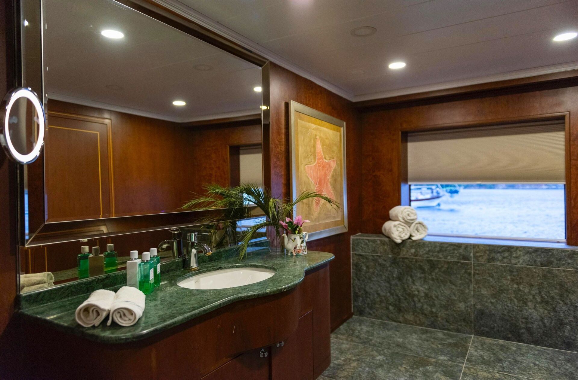 Luxurious bathroom with dark wood paneling, green marble countertop and tub, a large mirror, decorative plants, toiletries, rolled towels, starfish artwork, and a window overlooking water.