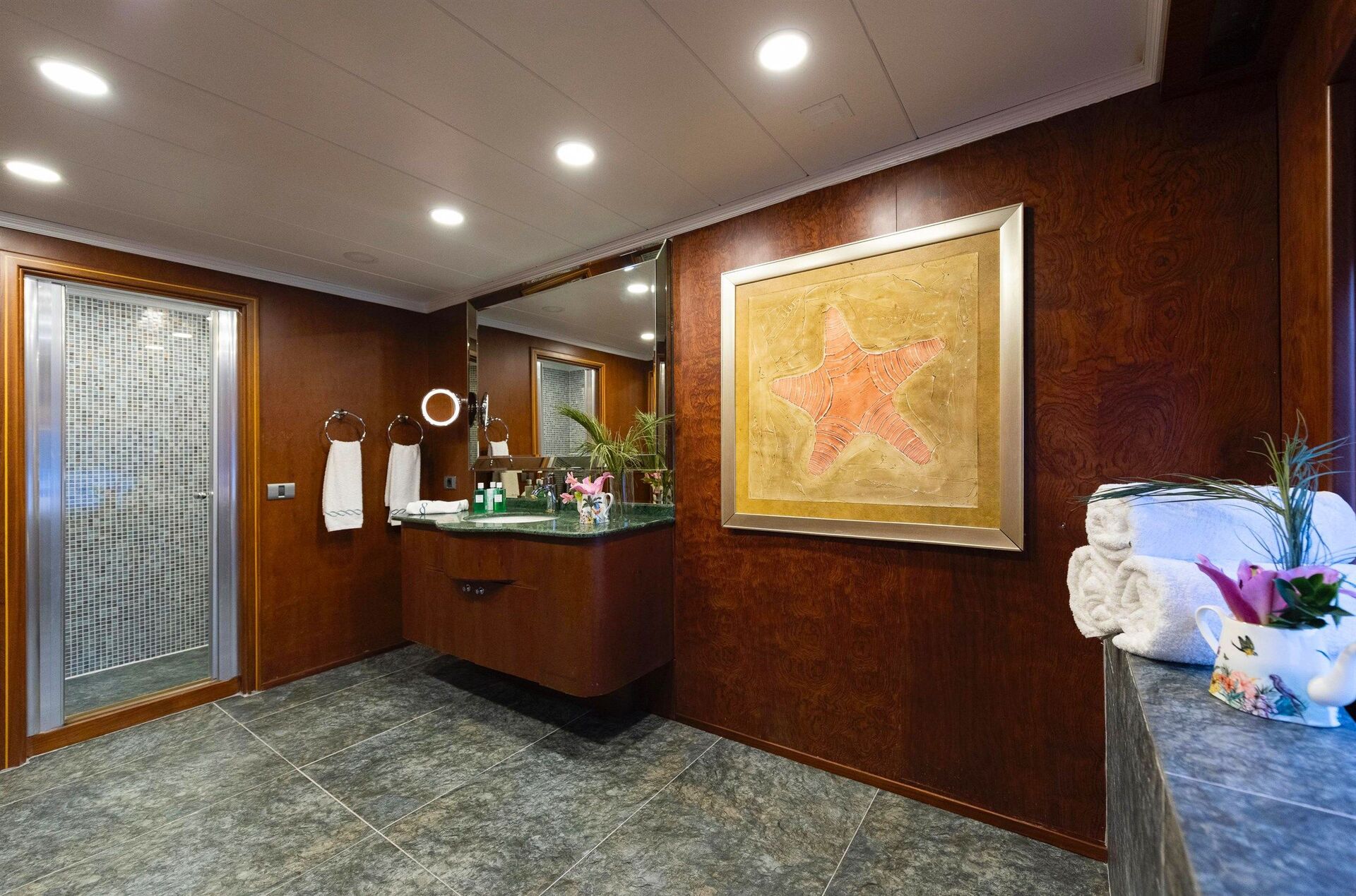 A spacious bathroom with dark wood walls, a large framed starfish artwork, a floating vanity with a mirror, towels, decorative plants, and a modern shower with glass door and mosaic tiles.