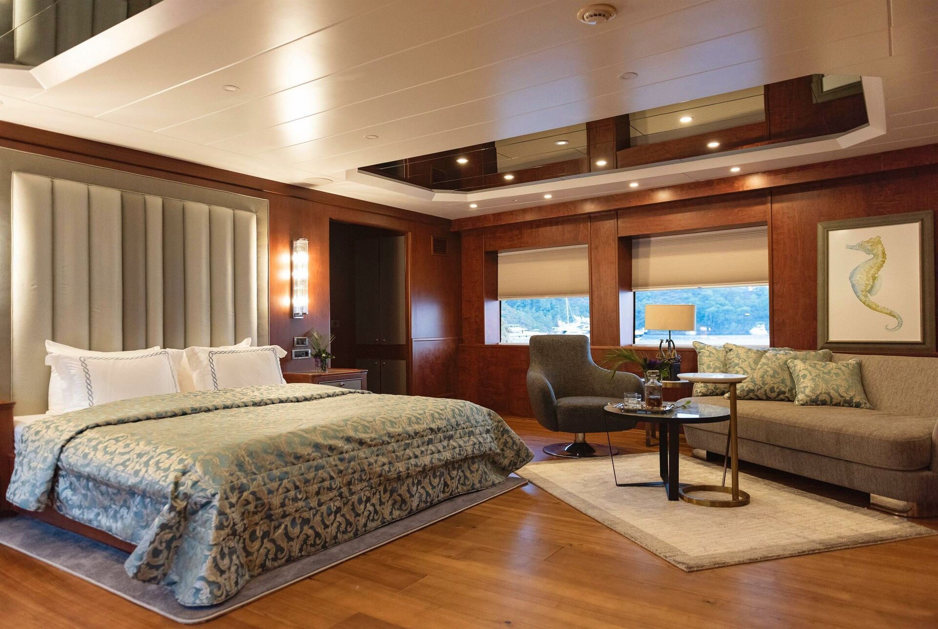 A spacious, elegant bedroom with a large upholstered bed, patterned bedding, wood-paneled walls, a gray sofa, a small table with chairs, a framed seahorse artwork, and large windows with shades.