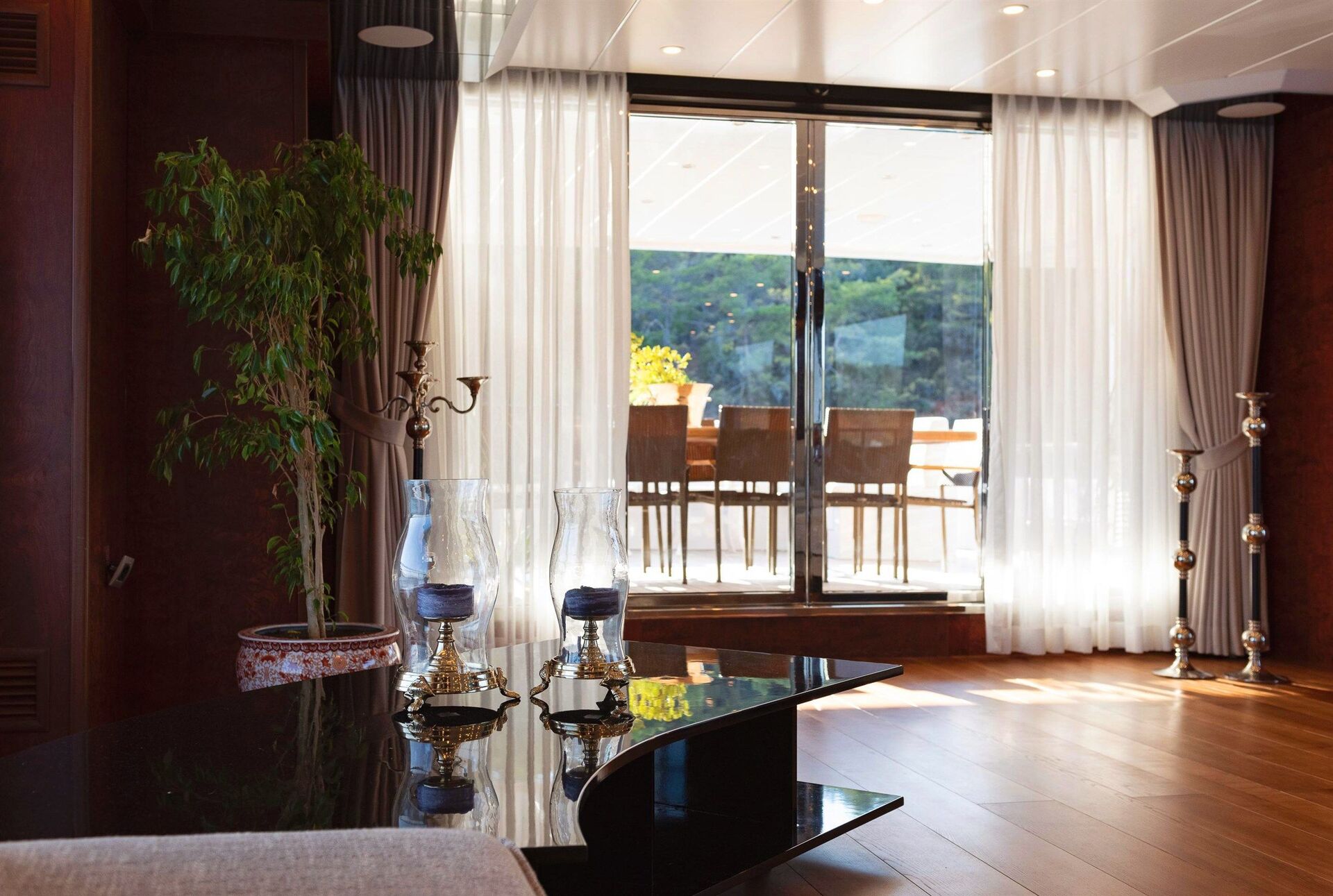 A bright, modern living room with wooden floors, a black glass coffee table, decorative candle holders, a potted plant, and large windows with sheer curtains opening to a sunny outdoor patio with chairs.