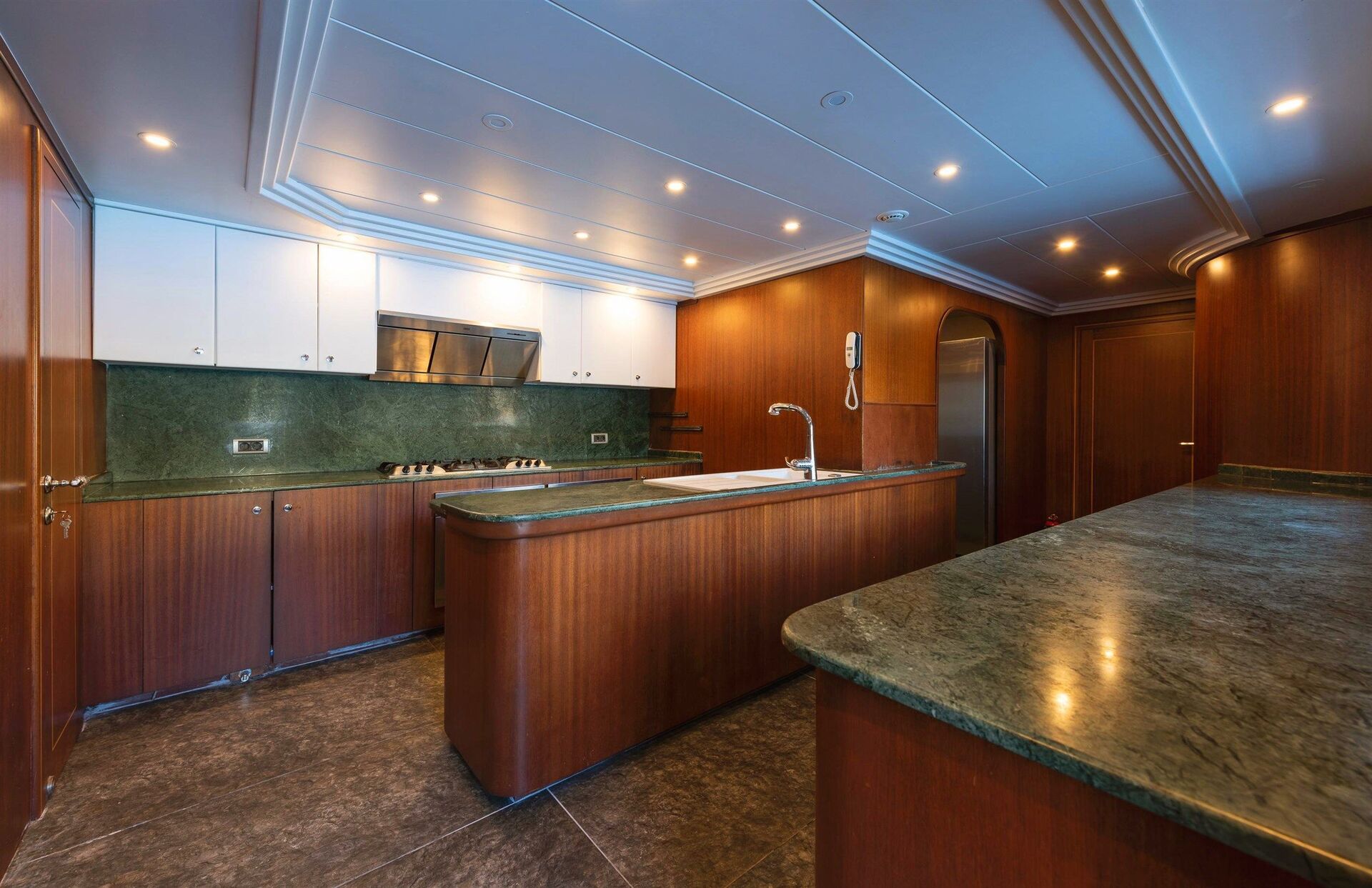 Modern kitchen with wood-paneled cabinets, green marble countertops, stainless steel appliances, under-cabinet lighting, and a central island. The floor is dark tile, and the ceiling has recessed lights.