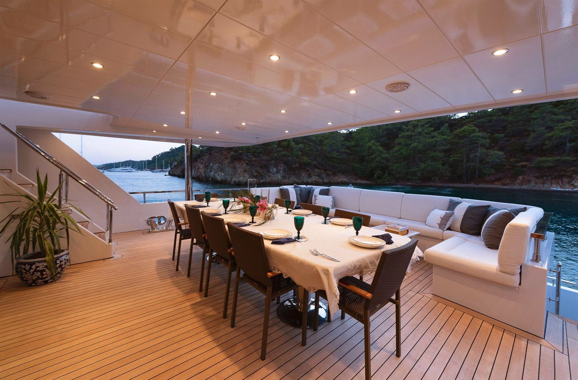 A luxurious yacht deck features a long dining table set for eight, surrounded by chairs and a white cushioned sofa. The deck overlooks calm water and a forested shoreline at sunset.