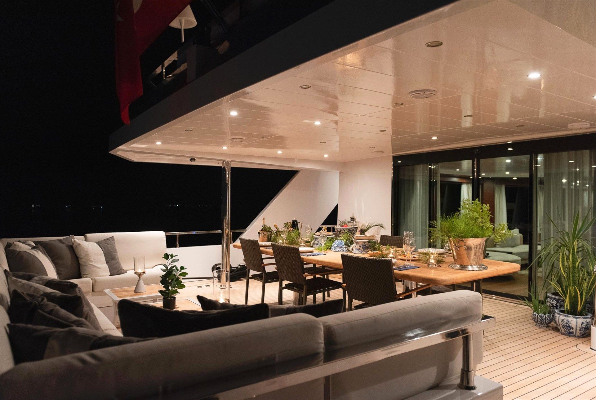 Elegant yacht deck at night with cozy seating area, dining table set for a meal, potted plants, and soft lighting, creating a warm and inviting atmosphere by large windows and glass doors.
