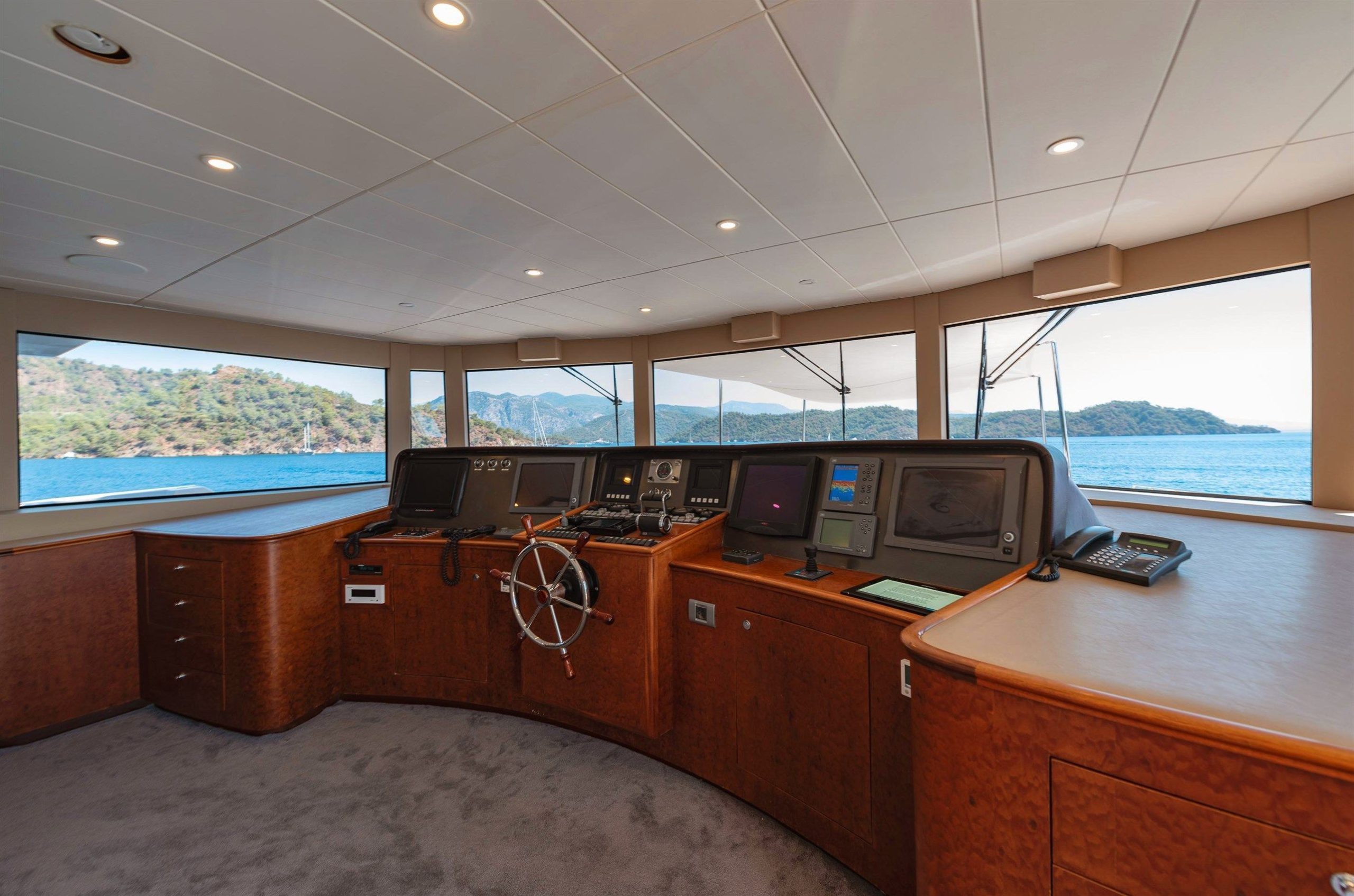 A spacious yacht bridge with large windows, wooden control panels, a steering wheel, multiple navigation screens, and a scenic view of the sea and distant green hills outside.