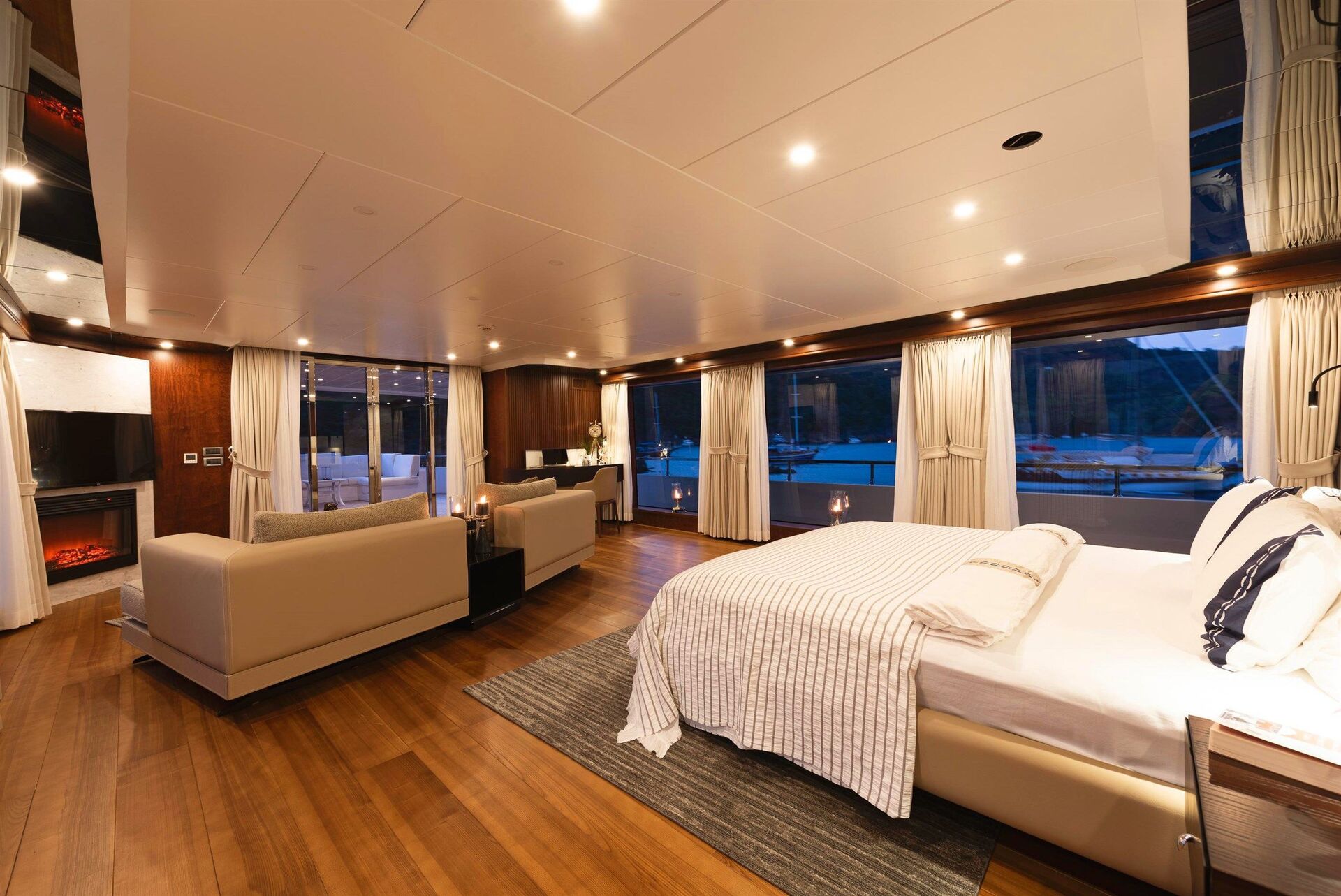 A spacious, modern bedroom with a large bed, cozy sitting area, wooden floors, and wide windows offering a view of a marina outside. Warm lighting and elegant decor create a luxurious, relaxing atmosphere.