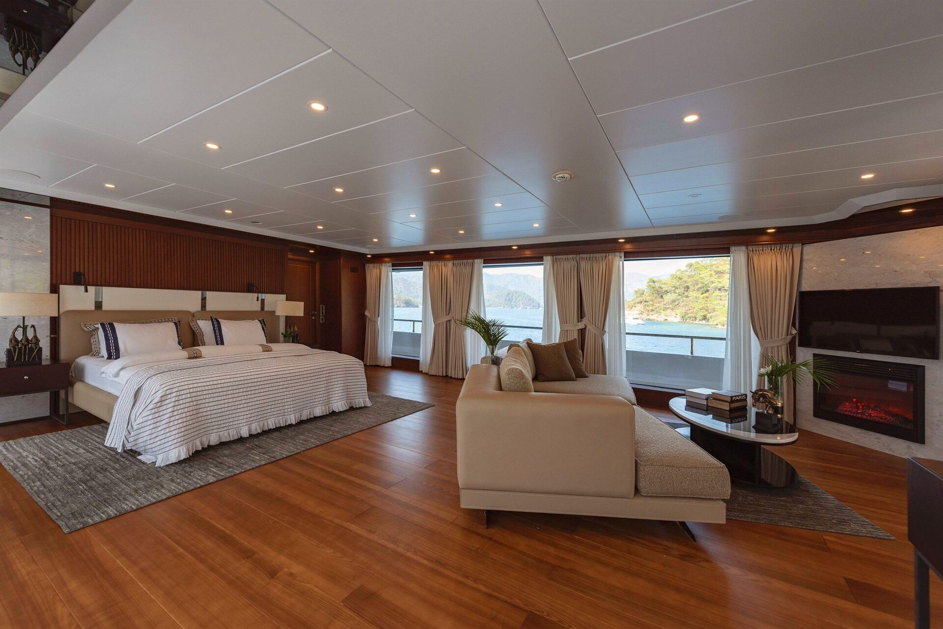 Spacious, modern yacht bedroom with a large bed, plush sofa, coffee table, TV, and electric fireplace. Floor-to-ceiling windows reveal scenic views of water and greenery outside.