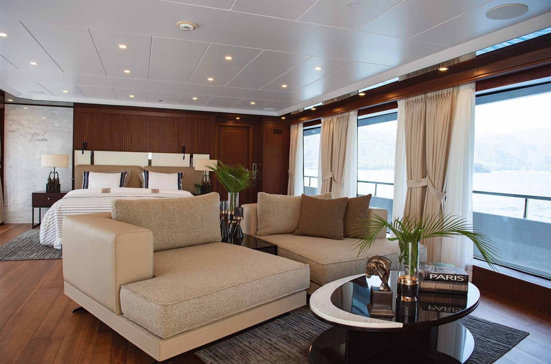 Luxurious yacht interior featuring a spacious bedroom with a large bed, modern beige sofas, a round coffee table with books and plants, large windows with curtains, and a view of water and mountains outside.