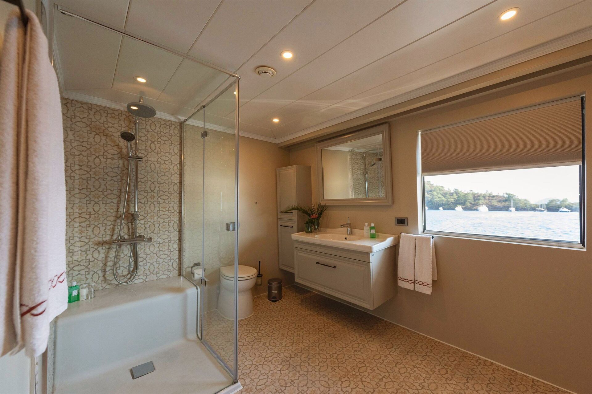 Modern bathroom with a glass shower, patterned tile walls and floors, a floating sink with a mirror above, towels, and a large window offering a scenic view of water and trees outside.