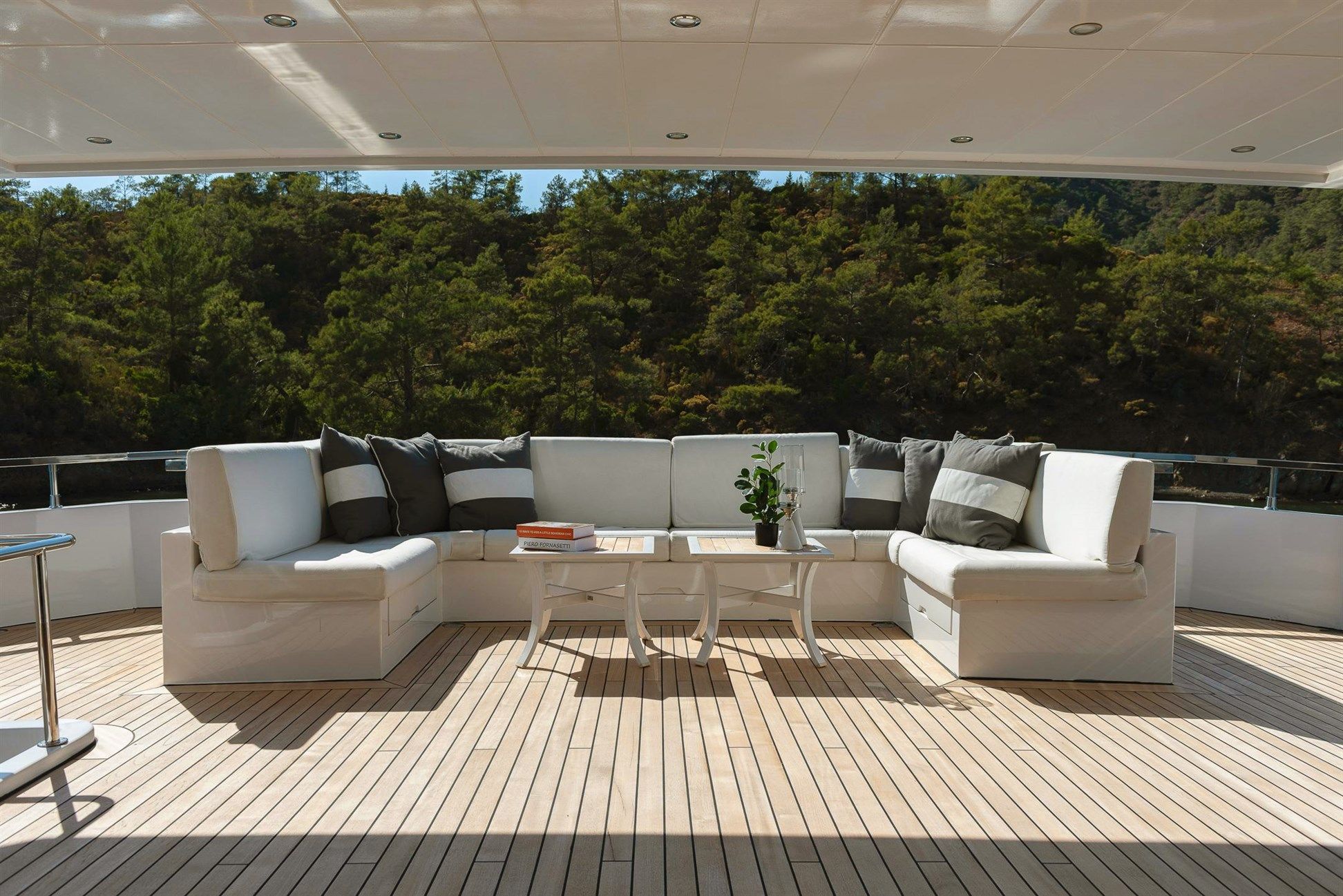 Luxurious outdoor seating area on a yacht with white cushioned sofas, gray and white pillows, a white table with books and a plant, and a view of green trees in the background.