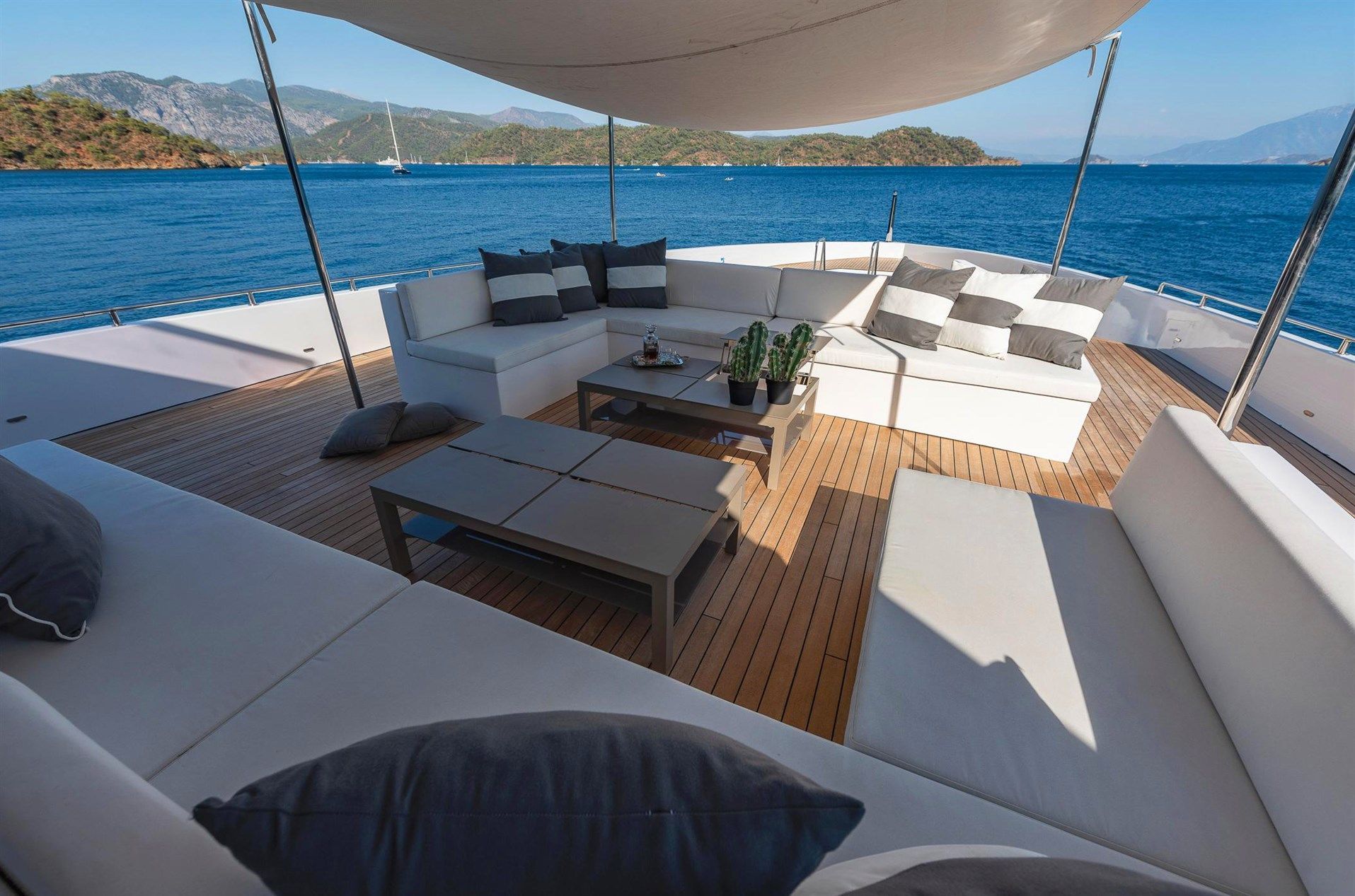 A luxurious yacht deck with white cushioned seating, gray pillows, wooden flooring, low coffee tables, and scenic views of blue sea and distant green islands under a shaded canopy.