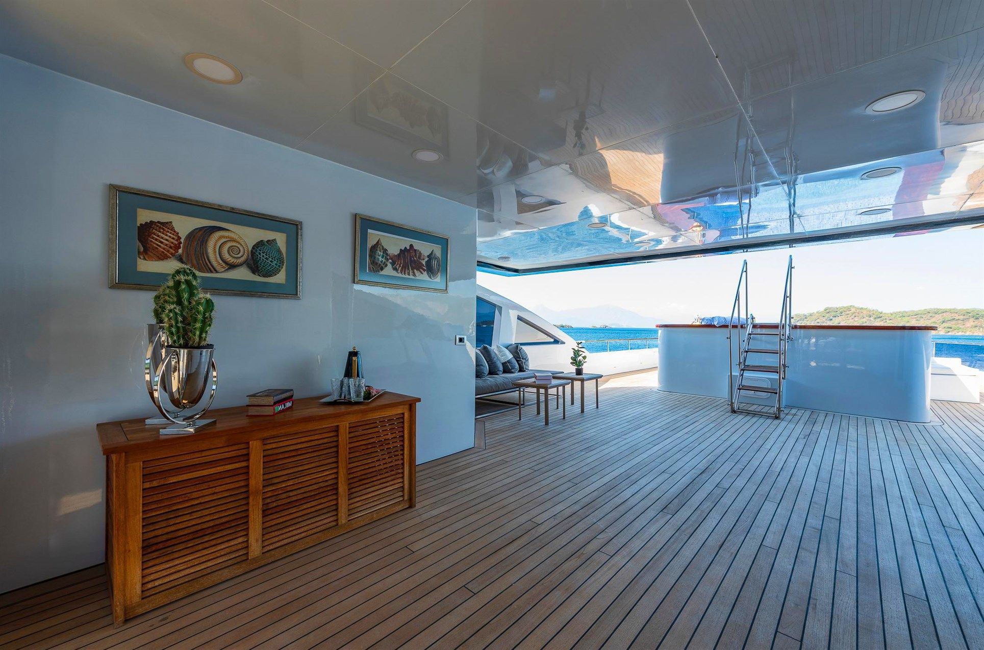 A spacious yacht deck with wooden flooring, a sideboard holding a cactus and champagne, two framed seashell artworks, lounge seating, and a ladder leading up, with ocean and islands visible in the background.