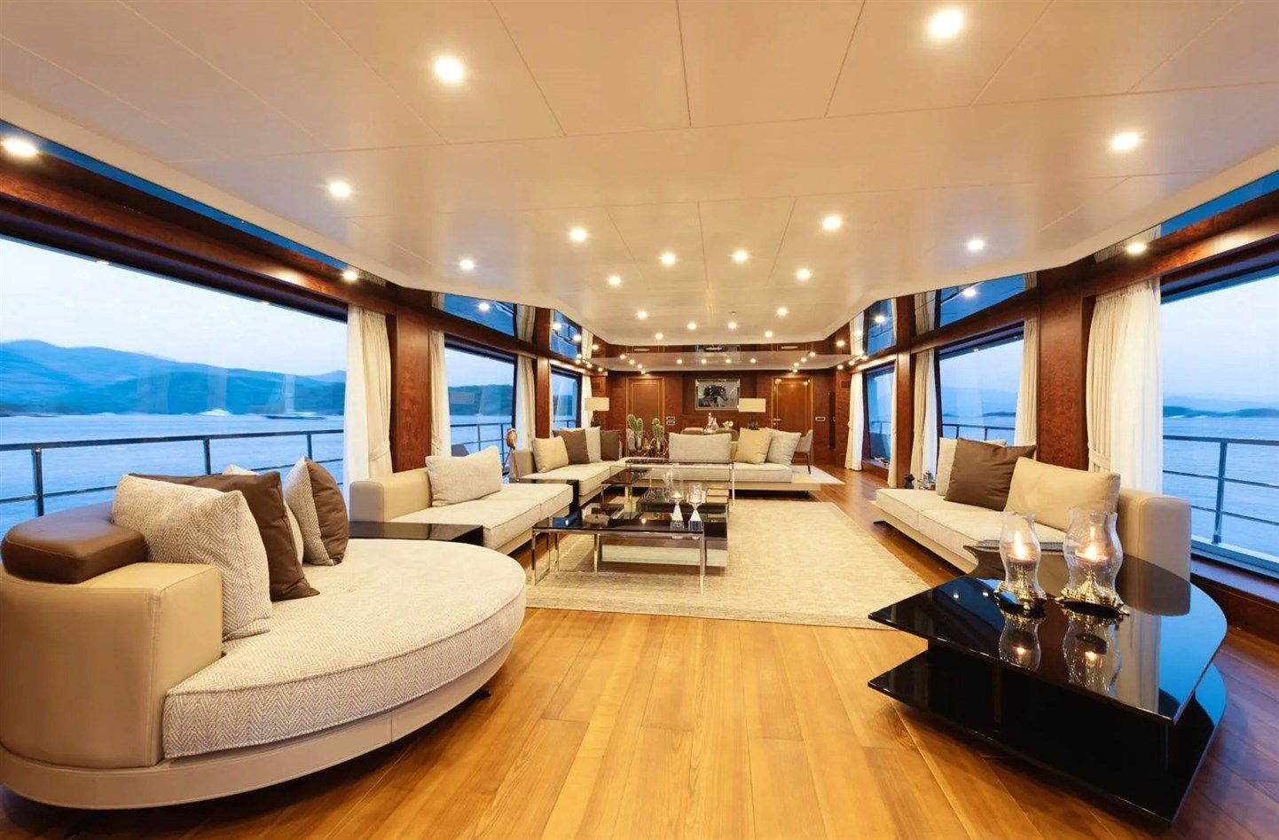 Spacious yacht lounge with large windows showing ocean views, modern sofas, a round beige sofa, wood floors, a glass coffee table, and soft ceiling lights. Decor includes cushions, candles, and small decorative items.