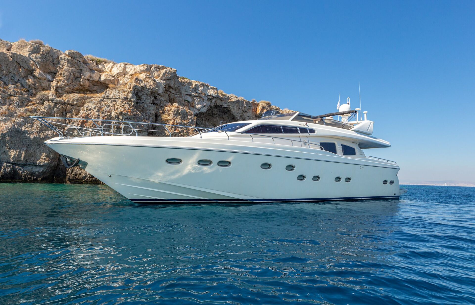 A white luxury yacht anchored near rocky cliffs on a clear, sunny day, with calm blue water and a bright blue sky in the background.
