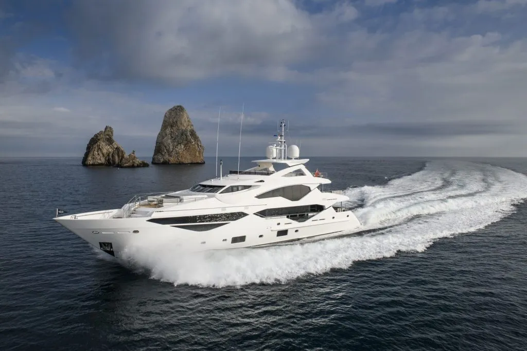 A large white luxury yacht speeds across calm blue water, leaving a wide wake behind it. Two tall rocky islands rise in the background under a partly cloudy sky.