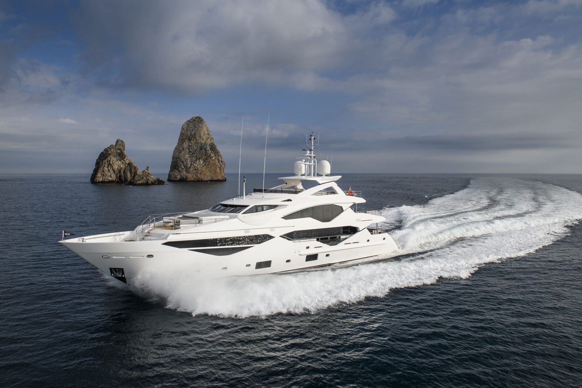 A large white luxury yacht speeds across calm blue water, leaving a wide wake behind it. Two tall rocky islands rise in the background under a partly cloudy sky.